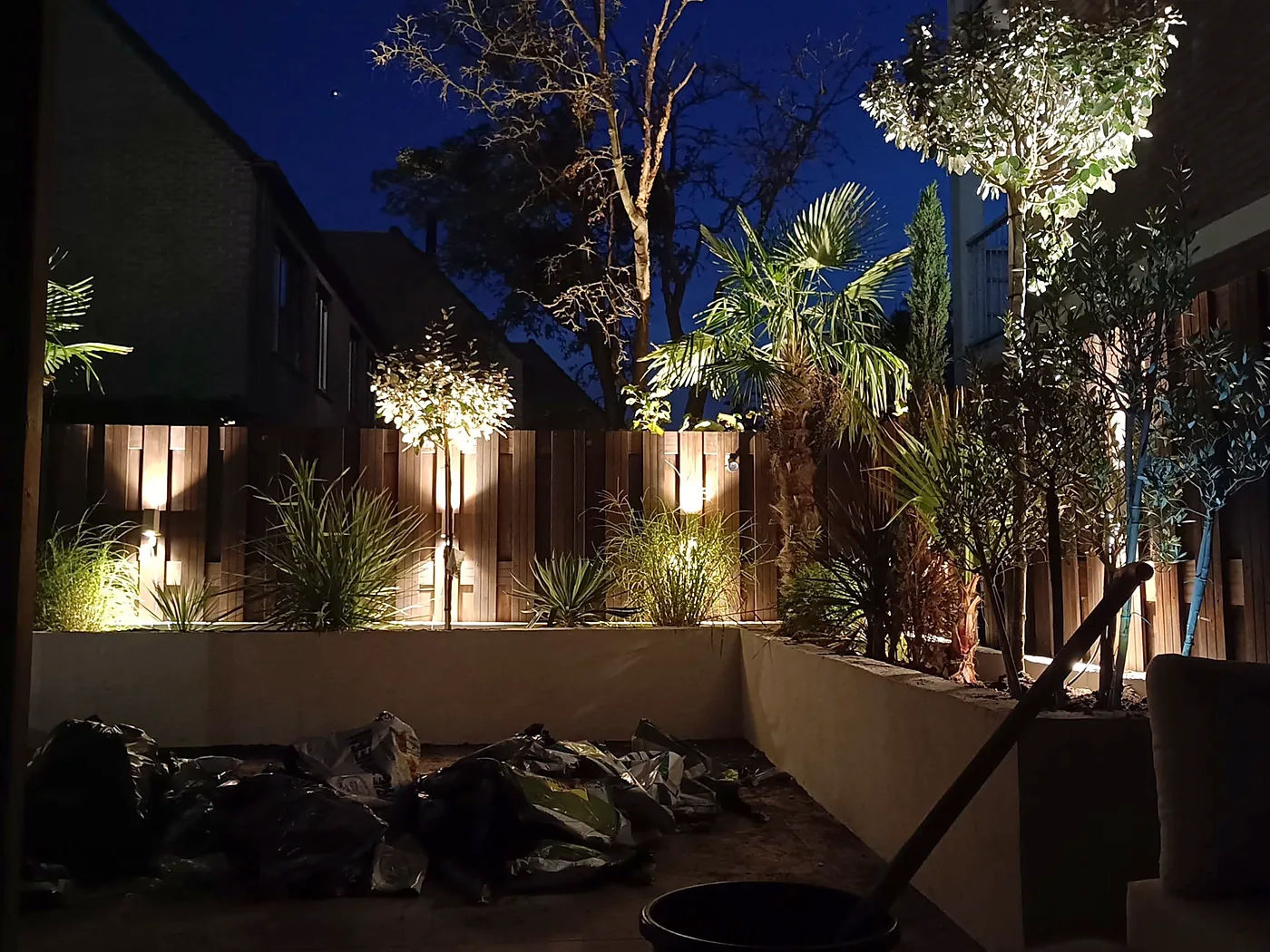 Dezelfde tuin 's avonds, verlicht met spots en wandlampen
